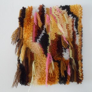 May include: A colorful, hand-woven wall hanging with a geometric pattern in shades of brown, orange, pink, and white. The hanging has a fringe border and is made with a variety of textures.