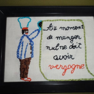 May include: Embroidered artwork featuring a chef holding a sign that reads "Au moment de manger nul ne doit avoir vergogne". The chef is wearing a blue and white uniform with a blue hat. The sign is green with black lettering.