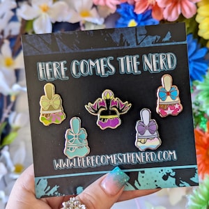 May include: A black card displaying five enamel pins with a cartoon design. The pins have a gold outline and are decorated with bows and colourful details. The text "HERE COMES THE NERD" and the website address are also visible.