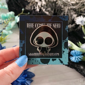 May include: A silver enamel pin featuring a cartoon character with a black hooded head and large eyes. The pin is on a black background with white text that reads "HERE COMES THE NERD" and a website address.