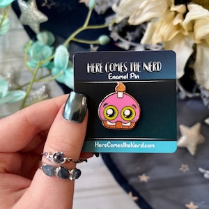 Op de afbeelding: Een roze cupcake emaille pin met een brandende kaars, grote ogen en een oranje mond. De pin zit op een kaart met "HERE COMES THE NERD" en "Enamel Pin". De website HereComesTheNerd.com is ook zichtbaar. Een leuk, kleurrijk accessoire.