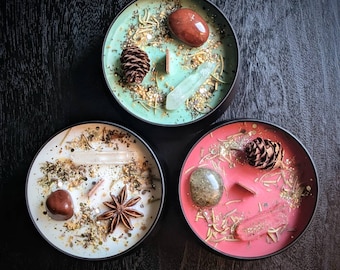 May include: Three round candles in black tins, each with a different color wax: mint green, white, and coral. The candles are decorated with crystals, pine cones, and dried herbs. The candles are on a dark wooden surface.