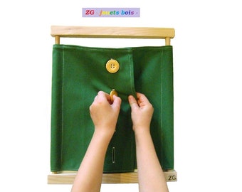 Può includere: Una cornice di legno con un pannello di tessuto verde. Due grandi bottoni di legno sono cuciti sul tessuto. Un bottone è chiuso e l'altro è in fase di chiusura da una mano.