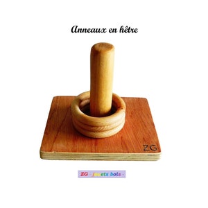 Puede incluir: Juguete de anillos de madera para apilar con una base y un poste central. Los anillos son de madera natural y tienen un color marrón claro. La base es de madera contrachapada y tiene un color marrón oscuro. El juguete está etiquetado con "ZG - jouets bois -" en la esquina inferior derecha.