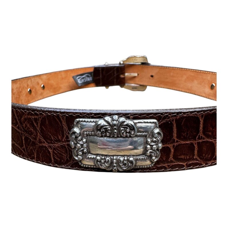 Może przedstawiać: Brązowy sk&oacute;rzany pasek o fakturze krokodylej. Pasek posiada prostokątną, srebrną klamrę z ozdobnymi detalami. Klamra jest centralnym punktem, dodając odrobinę elegancji do designu paska.