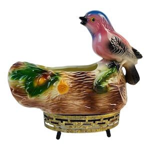 May include: A ceramic planter shaped like a tree log with a pink and blue bird perched on top. The planter has a metal stand with three legs and is decorated with green leaves and acorns.