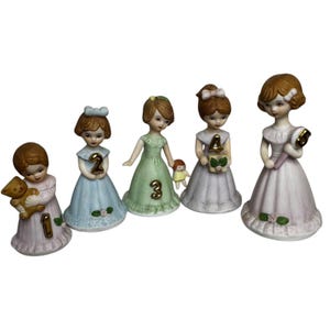 May include: Five porcelain figurines of young girls in pastel dresses. Each girl holds a different item, including a teddy bear, a number, and a doll. These figurines are likely intended as birthday cake toppers.