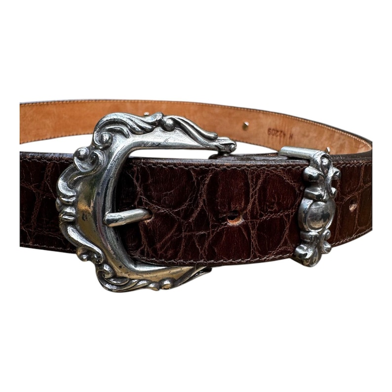Może przedstawiać: Brązowy sk&oacute;rzany pasek ze srebrną, ozdobną klamrą. Pasek ma teksturowany wz&oacute;r przypominający krokodyla. Klamra to duży, dekoracyjny element o zakrzywionym kształcie. Klasyczny dodatek.