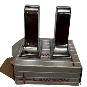 Pode incluir: Dois acessórios de casa de banho cromados, possivelmente toalheiros, são exibidos numa caixa listrada cinza e branca. A caixa tem o nome da marca "LAWSON" impresso.