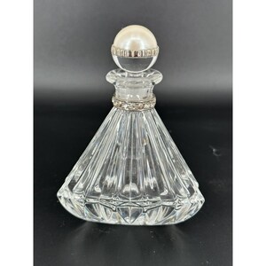 Pode incluir: Um frasco de perfume de vidro transparente com uma rolha de bola branca perolada. O frasco tem uma forma cónica facetada com uma faixa prateada adornada com pequenos cristais transparentes à volta do pescoço. A rolha é encimada por uma faixa de metal prateado.