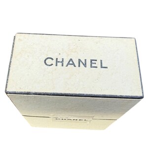 Op de afbeelding: Een rechthoekige, gebroken witte doos met het woord "CHANEL" in zwarte letters op de bovenkant. De doos lijkt van karton of een vergelijkbaar materiaal te zijn. Het ontwerp is eenvoudig en elegant.