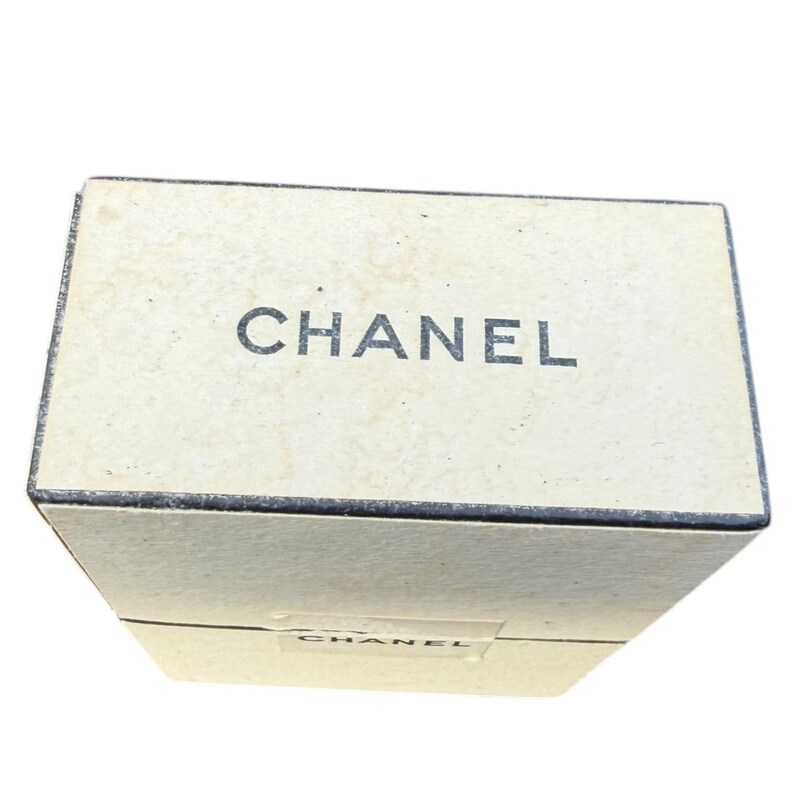 Op de afbeelding: Een rechthoekige, gebroken witte doos met het woord "CHANEL" in zwarte letters op de bovenkant. De doos lijkt van karton of een vergelijkbaar materiaal te zijn. Het ontwerp is eenvoudig en elegant.