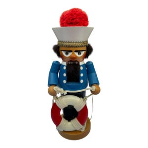 May include: A wooden sailor nutcracker with a blue jacket, white hat, and red pom-pom. The nutcracker holds a white and red life preserver with a rope. The sailor has a black beard and is standing on a wooden base.
