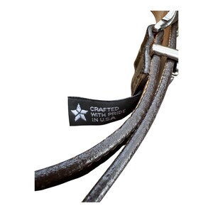 Może przedstawiać: Zbliżenie brązowego sk&oacute;rzanego paska ze srebrną klamrą. Czarna metka z białym tekstem i logo gwiazdy głosi "Crafted with Pride in U.S.A."