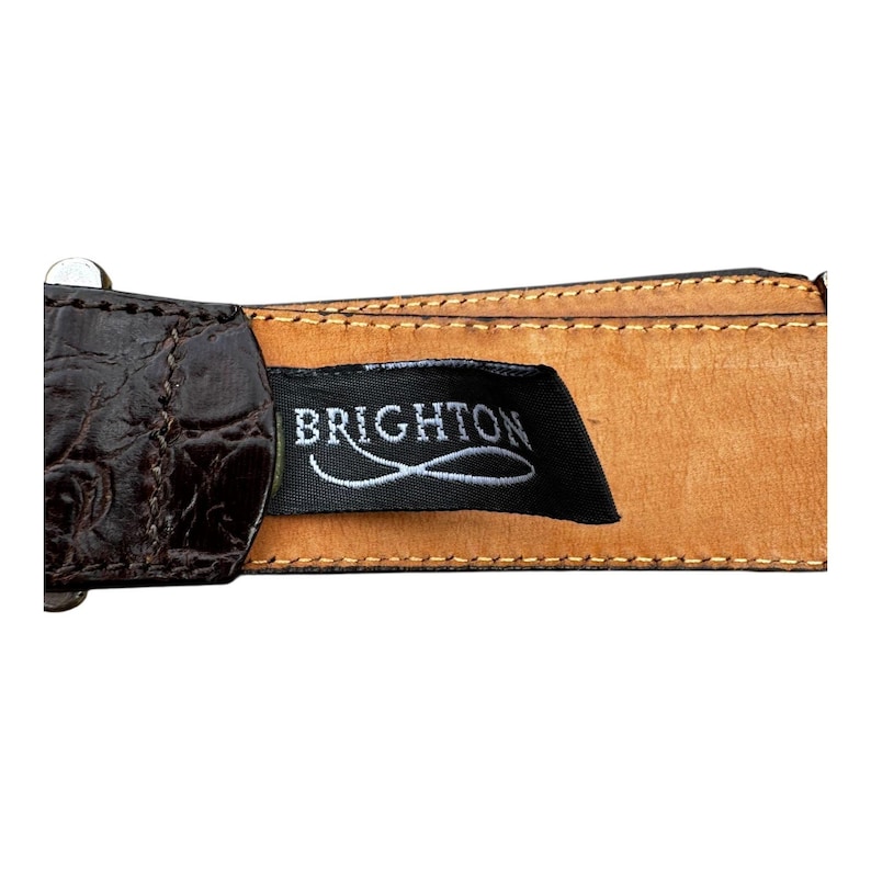 Może przedstawiać: Zbliżenie brązowego sk&oacute;rzanego paska z teksturowanym wzorem przypominającym krokodyla. Pasek posiada czarną metkę z nazwą marki "BRIGHTON" w białym skrypcie. Pasek jest przeszyty jasnobrązową nicią.