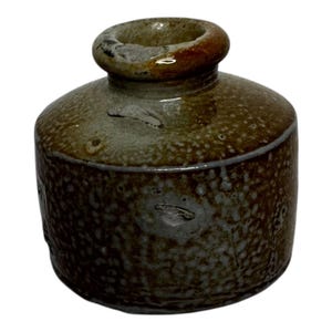 May include: A small, handcrafted ceramic bottle with a rounded body and a short, cylindrical neck. The bottle has a speckled, mottled appearance with shades of brown, gray, and tan. The opening is a darker brown color.