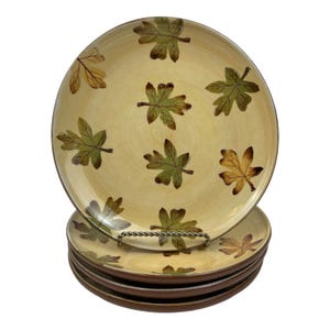 May include: A stack of four round ceramic plates with a cream-colored base and a hand-painted pattern of scattered autumn leaves in shades of green, yellow, and brown. The plates feature a smooth, glossy finish and a dark brown rim.