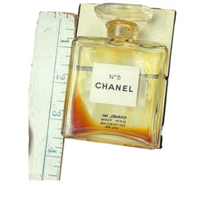 Op de afbeelding: Een vintage Chanel No. 5 parfumflesje met een doorzichtige glazen stopper. De fles is rechthoekig met een wit etiket met de tekst "N&deg;5 CHANEL". Een vintage liniaal ligt naast de fles. De parfum binnenin is amberkleurig.