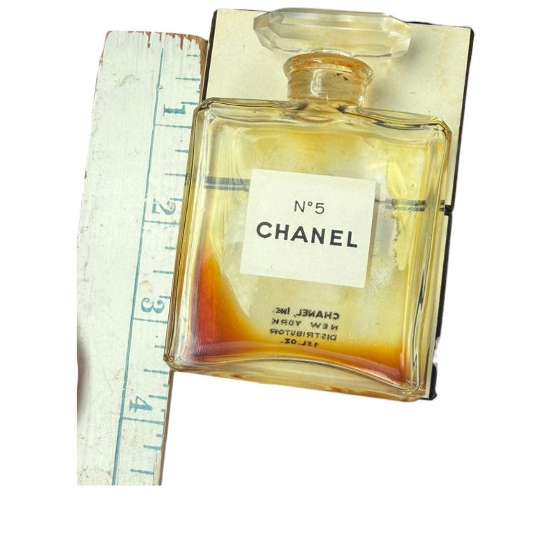 Op de afbeelding: Een vintage Chanel No. 5 parfumflesje met een doorzichtige glazen stopper. De fles is rechthoekig met een wit etiket met de tekst "N&deg;5 CHANEL". Een vintage liniaal ligt naast de fles. De parfum binnenin is amberkleurig.