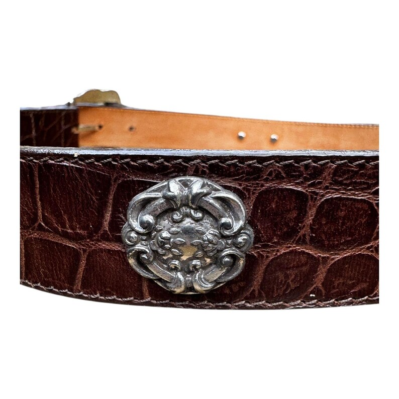 Może przedstawiać: Zbliżenie brązowego sk&oacute;rzanego paska ze srebrną ozdobną klamrą. Pasek ma fakturę przypominającą sk&oacute;rę krokodyla. Klamra jest ozdobiona kwiatowym wzorem.