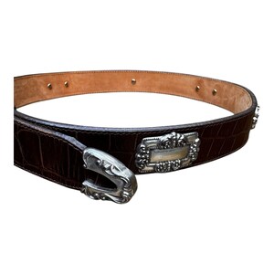 Może przedstawiać: Brązowy sk&oacute;rzany pasek o fakturze krokodylej. Pasek posiada srebrną klamrę i ozdobną srebrną płytkę. Klasyczny dodatek.