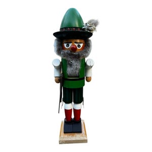 Könnte beinhalten: Holz-Nussknacker-Figur in traditionellem deutschem Design. Die Figur hat einen grünen Hut, eine Jacke und eine Hose mit einem Bart und Schnurrbart aus Kunstfell. Der Nussknacker ist ca. 30 cm hoch.