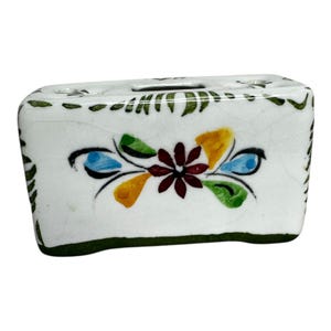 Puede incluir: Un portacepillos rectangular de cerámica blanca con un diseño floral. El soporte presenta una flor central con pétalos granates, rodeada de detalles azules, amarillos y verdes. Los bordes superior e inferior están bordeados con adornos verdes y negros.
