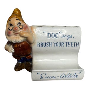 May include: A ceramic toothbrush holder featuring a cartoon character, Doc, from Snow White. The holder is white with blue text that reads "DOC says, BRUSH YOUR TEETH" and "Snow-White". The character is wearing a brown coat, blue pants, and a yellow hat.
