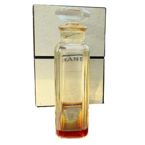 Op de afbeelding: Een vintage parfumflesje met een helder, rechthoekig ontwerp en een facetgeslepen stop. De fles heeft een licht amberkleurige tint met een rode basis en het woord "HANE" is op de voorkant gedrukt. De fles staat tegen een cr&egrave;mekleurige doos.