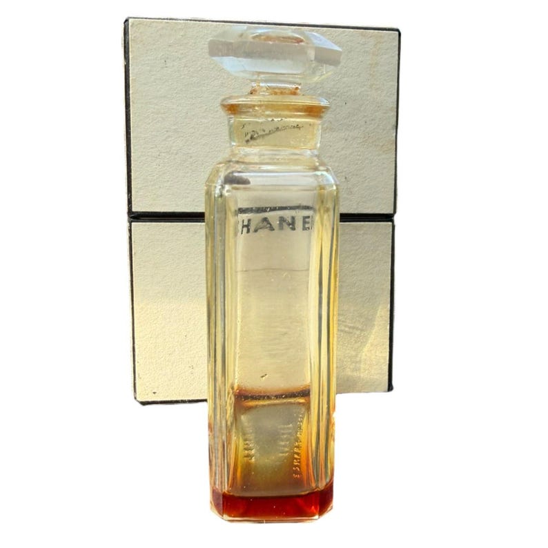 Op de afbeelding: Een vintage parfumflesje met een helder, rechthoekig ontwerp en een facetgeslepen stop. De fles heeft een licht amberkleurige tint met een rode basis en het woord "HANE" is op de voorkant gedrukt. De fles staat tegen een cr&egrave;mekleurige doos.