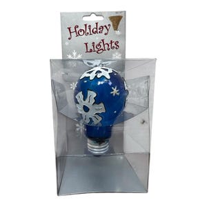 Op de afbeelding: Een blauwe kerstlamp met witte sneeuwvlokontwerpen, verpakt in een transparante plastic doos. De verpakking bevat de tekst "Holiday Lights" in rode letters.