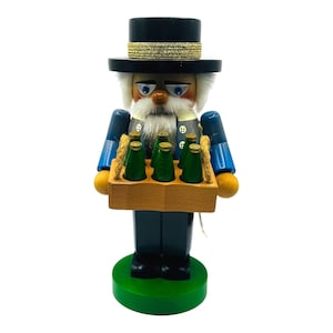 May include: Wooden nutcracker figurine of a man in a black top hat with a gold band, blue coat, and a white beard. The figurine holds a wooden tray with six green bottles. The base is green.