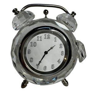 Puede incluir: Un reloj despertador de cristal transparente y facetado con un marco de metal plateado. La esfera del reloj es blanca con números y manecillas negras. El reloj tiene un asa curva y dos alarmas en forma de campana de cristal.