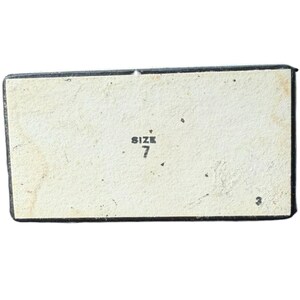 Op de afbeelding: Rechthoekige doos met een lichtbeige binnenkant en een zwarte rand. De tekst "SIZE 7" is zwart afgedrukt, gecentreerd aan de binnenkant. De doos heeft een versleten, vintage uitstraling.