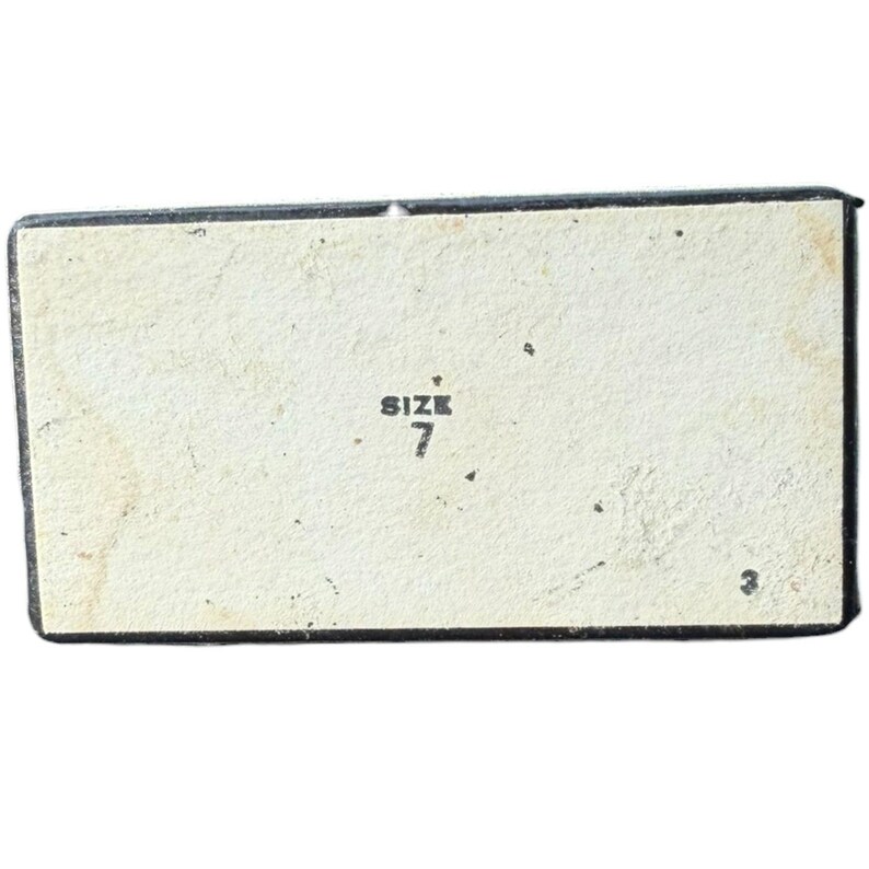 Op de afbeelding: Rechthoekige doos met een lichtbeige binnenkant en een zwarte rand. De tekst "SIZE 7" is zwart afgedrukt, gecentreerd aan de binnenkant. De doos heeft een versleten, vintage uitstraling.