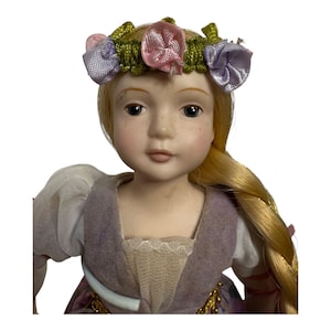 Pode incluir: Boneca de porcelana com cabelo loiro e olhos azuis, vestindo um vestido lavanda e uma coroa de flores com detalhes rosa e roxo. A boneca tem sardas e uma expressão serena, com uma longa trança.