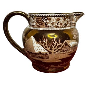 May include: A brown and gold ceramic pitcher with a handle and spout. The pitcher features a winter landscape scene with a cabin, trees, and a mountain. The rim is decorated with floral patterns.