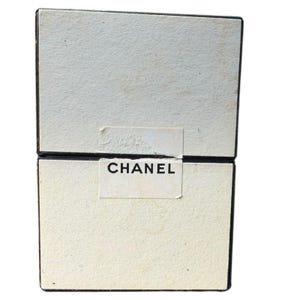Op de afbeelding: Twee gestapelde, gebroken witte dozen met zwarte rand. Het woord "CHANEL" is in zwart gedrukt op een wit etiket dat op de voorkant van de dozen is aangebracht. De dozen hebben een getextureerd oppervlak.