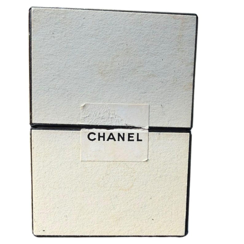 Op de afbeelding: Twee gestapelde, gebroken witte dozen met zwarte rand. Het woord "CHANEL" is in zwart gedrukt op een wit etiket dat op de voorkant van de dozen is aangebracht. De dozen hebben een getextureerd oppervlak.