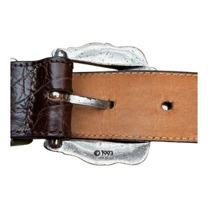 Może przedstawiać: Zbliżenie brązowego sk&oacute;rzanego paska ze srebrną klamrą. Pasek ma teksturowany wz&oacute;r przypominający krokodyla na ciemnobrązowej sk&oacute;rze. Klamra jest ozdobna z dekoracyjnym wzorem, a rok "1993" jest wytłoczony na klamrze.