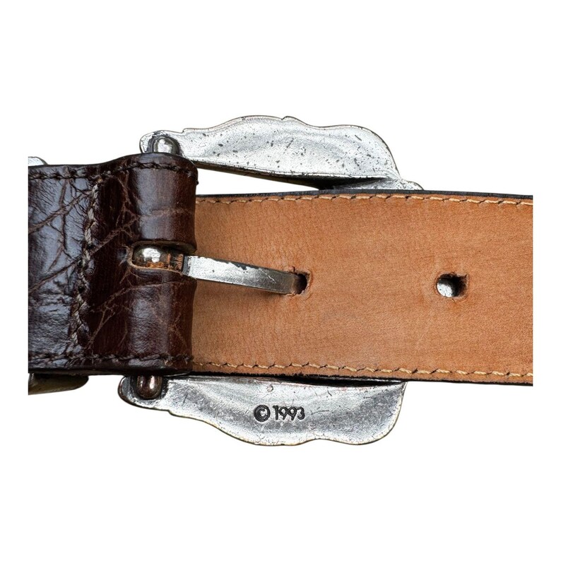 Może przedstawiać: Zbliżenie brązowego sk&oacute;rzanego paska ze srebrną klamrą. Pasek ma teksturowany wz&oacute;r przypominający krokodyla na ciemnobrązowej sk&oacute;rze. Klamra jest ozdobna z dekoracyjnym wzorem, a rok "1993" jest wytłoczony na klamrze.