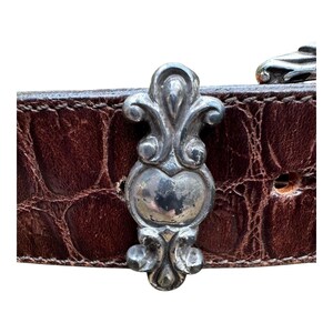 Może przedstawiać: Zbliżenie brązowego sk&oacute;rzanego paska o fakturze krokodyla. Pasek posiada ozdobną srebrną klamrę o bogatym wzornictwie. Klamra jest wyśrodkowana na pasku, prezentując jego skomplikowane detale.