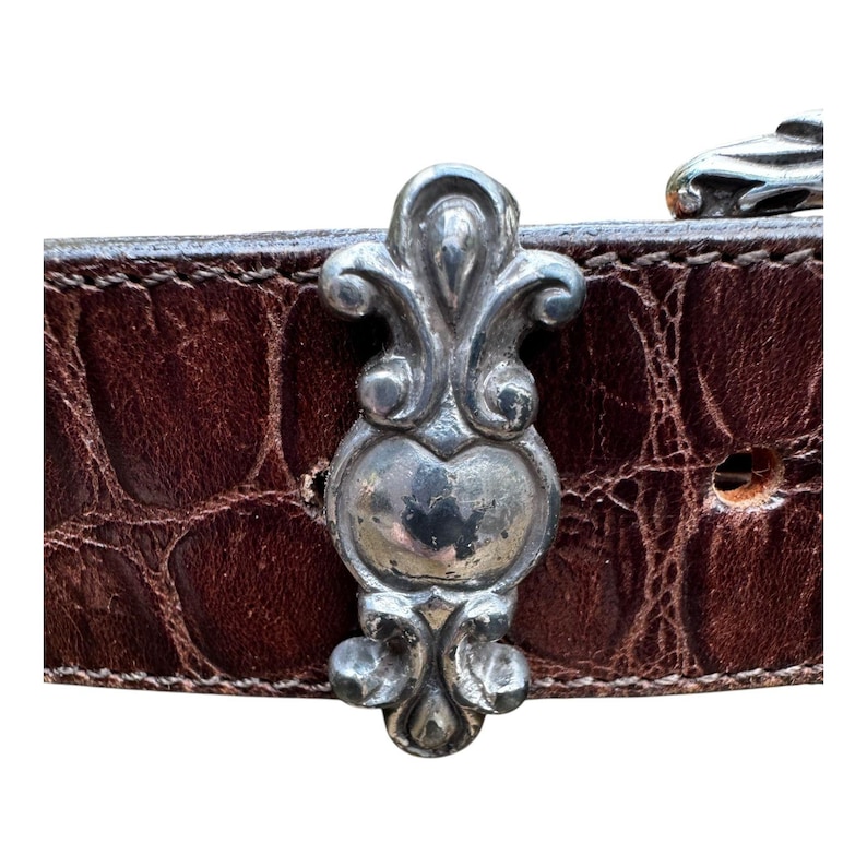 Może przedstawiać: Zbliżenie brązowego sk&oacute;rzanego paska o fakturze krokodyla. Pasek posiada ozdobną srebrną klamrę o bogatym wzornictwie. Klamra jest wyśrodkowana na pasku, prezentując jego skomplikowane detale.