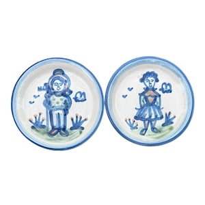 Puede incluir: Dos platos redondos de cerámica con un diseño azul y blanco. Cada plato presenta una ilustración pintada a mano de una persona con atuendo azul, rodeada de motivos florales y de nubes estilizados. Los platos tienen un borde azul.