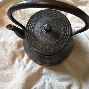 May include: A black cast iron teapot with a curved handle and a spout. The lid features a circular design. The teapot is sitting on a cream-colored fabric.