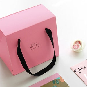 May include: A pink gift bag with black handles and the text "MERCI BEAUCOUP" printed on the front. The bag is sitting on a white surface next to a pink card with the word "love" written on it.