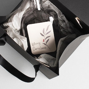 Op de afbeelding: Een zwarte cadeauzak met een zwarte lintgreep. De zak is open en bevat een heldere glazen fles die is ingepakt in wit tissuepapier. Een klein kaartje met de tekst "Prettige Kerst" en een eenvoudig bladontwerp is zichtbaar.