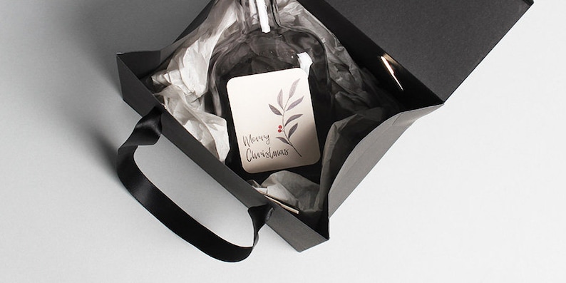 Op de afbeelding: Een zwarte cadeauzak met een zwarte lintgreep. De zak is open en bevat een heldere glazen fles die is ingepakt in wit tissuepapier. Een klein kaartje met de tekst "Prettige Kerst" en een eenvoudig bladontwerp is zichtbaar.