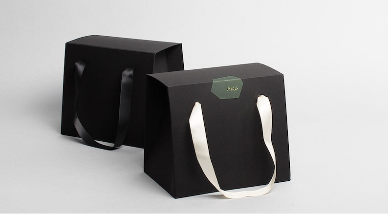 Op de afbeelding: Twee zwarte cadeaudozen met handvatten. E&eacute;n doos heeft een zwart lint handvat, en de andere heeft een wit lint handvat. De doos met het witte lint handvat heeft een groene sticker met gouden letters die "Laid." zeggen.