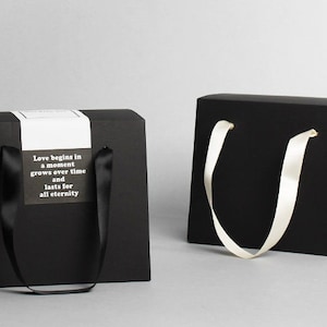 Op de afbeelding: Twee zwarte cadeauzakken met handvatten. E&eacute;n tas heeft een wit label met de tekst "Liefde begint in een moment, groeit met de tijd en duurt voor altijd".